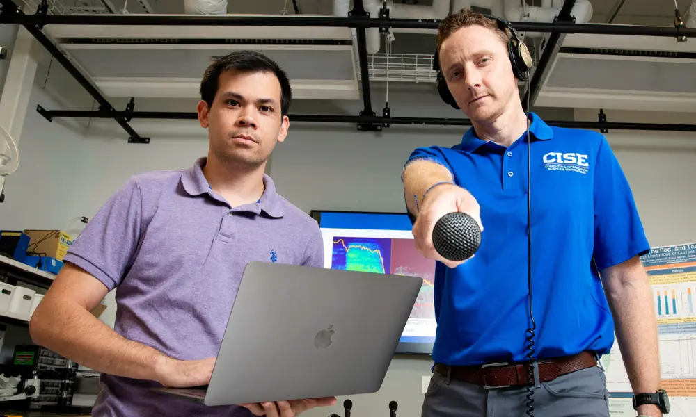 Men staring at camera - one is holding a microphone toward camera while the other one is holding a computer